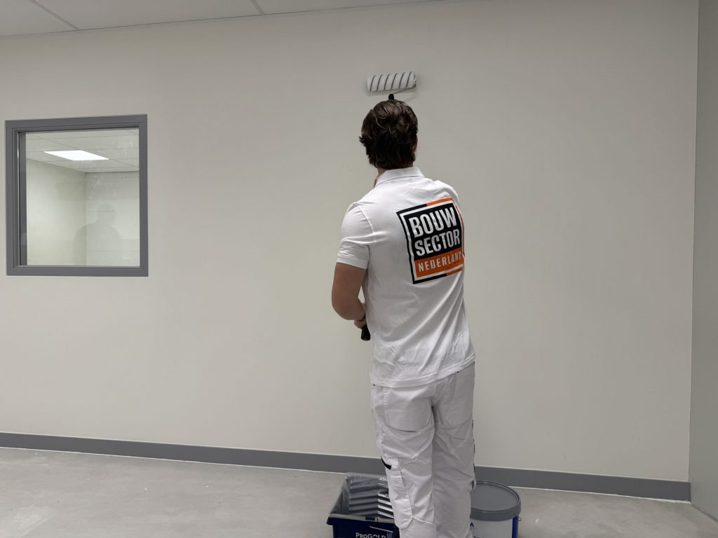 Schilder in wit werktenue saust een lichte wand, met een verfbak en een emmer muurverf aan zijn voeten