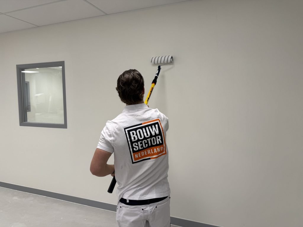 Schilder rolt een witte wand met een verlengsteel, naast een binnenraam met grijs kozijn