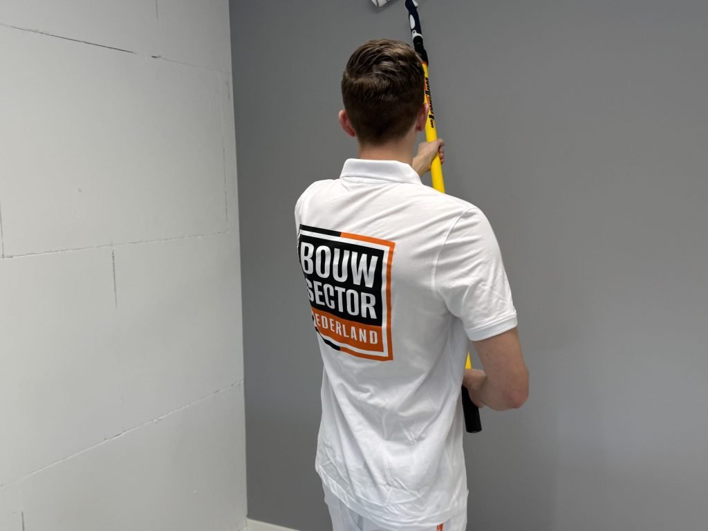 Schilder met logo van Bouwsector Nederland rolt met een lange steel een grijze wand naast een kale kalkzandsteenmuur