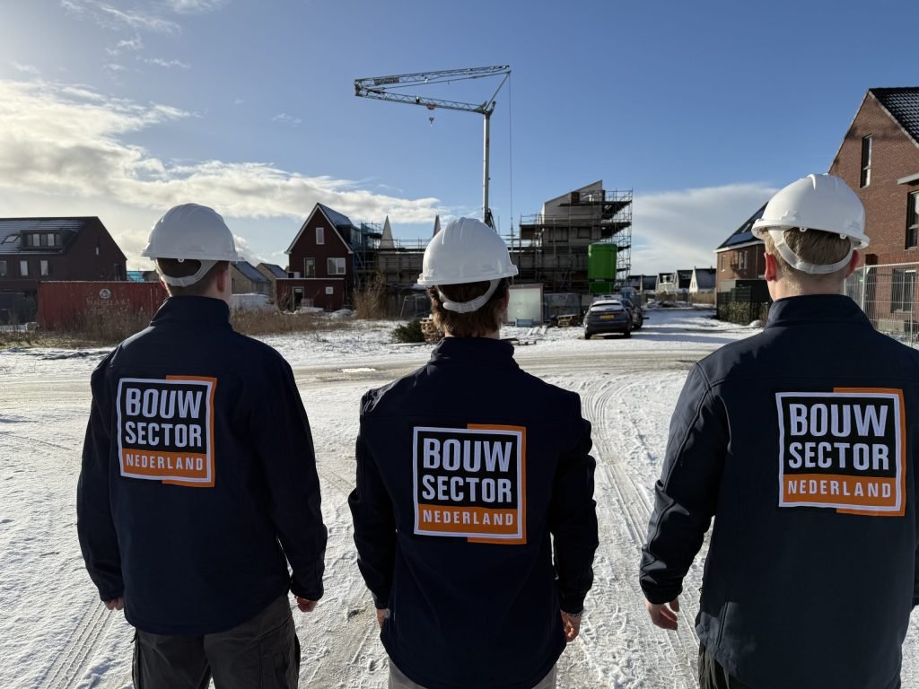 Drie medewerkers van Bouwsector Nederland in donkerblauwe jas en met bouwhelm kijken naar een nieuwbouwstraat in de sneeuw